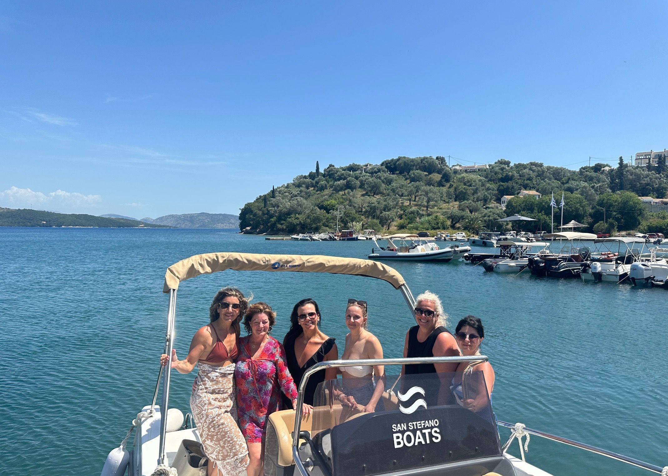 Six retreat guests on a small boat in a calm blue bay with olive-covered hills behind