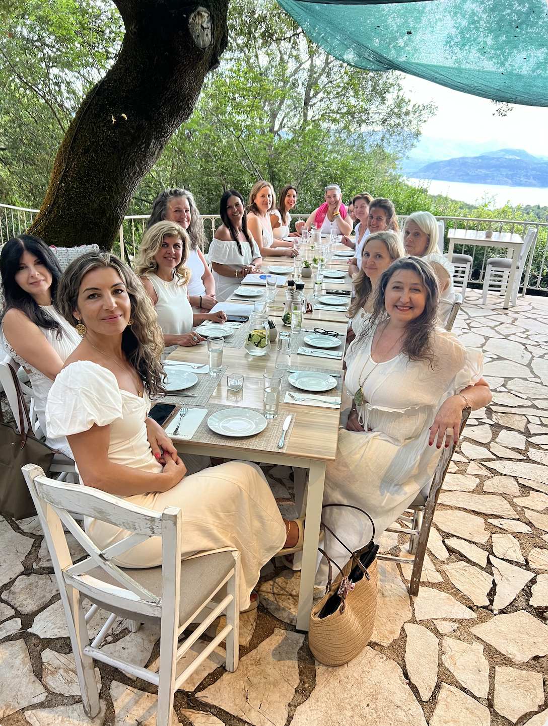 Retreat guests dressed in white around a long table beneath an old tree, with the sea and mountains beyond